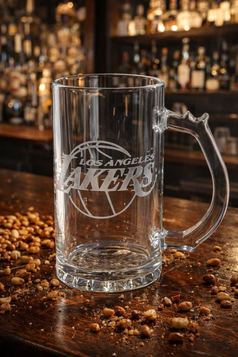 Clear glass mug with 'Los Angeles Lakers' engraving on a wooden bar surface with scattered coffee beans.